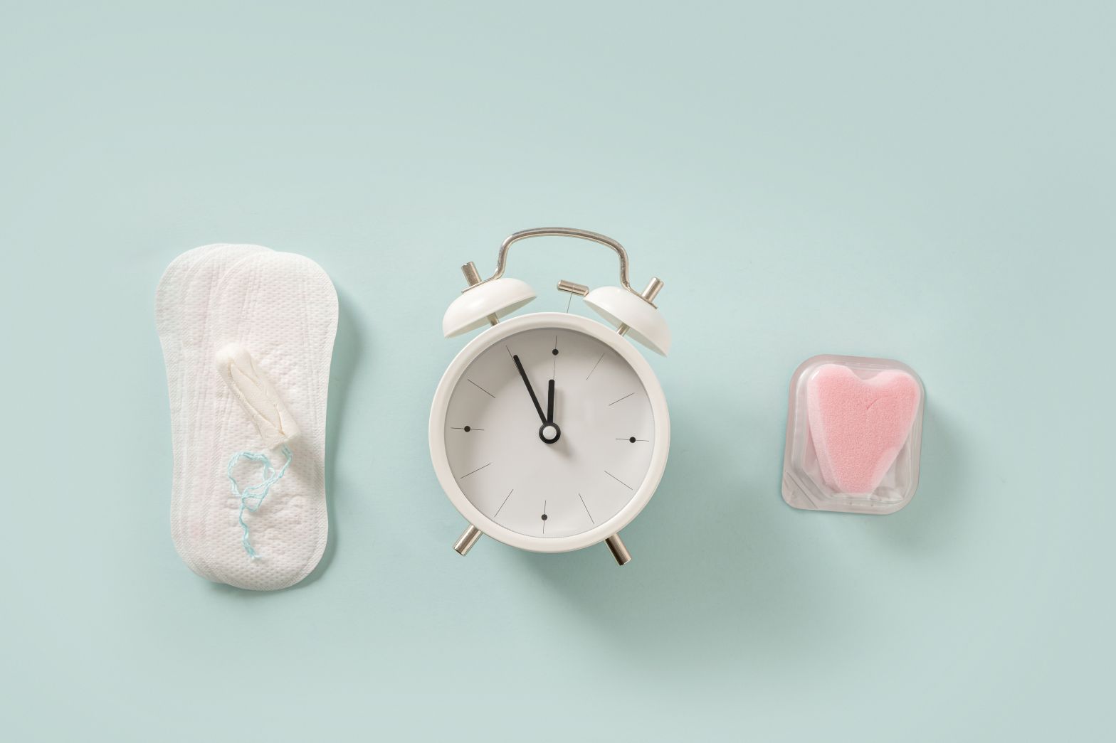 Menstrual products and birth control sponge alongside an analog alarm clock on a light blue background